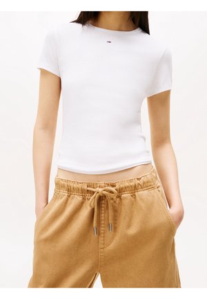 Vrouw draagt een aansluitende witte korte mouw crop top en losse beige broek met trekkoorden, handen in de zakken.