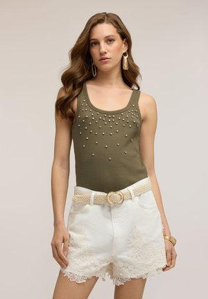 Femme portant un haut sans manches vert olive avec de petites perles et un short blanc orné de dentelle, accessoirisé avec une ceinture tressée et des bijoux en or.