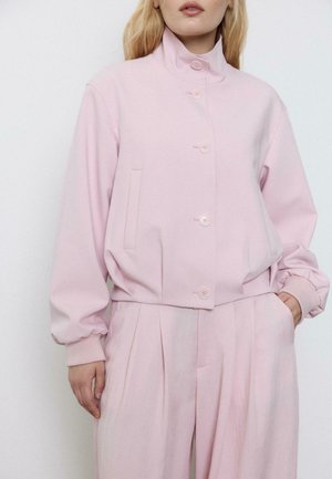 Žena v světle růžovém zapínacím kabátku s vysokým límcem a sladěných volně střižených plisovaných kalhotách, ruce v kapsách.