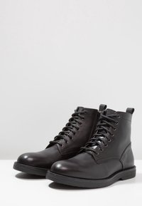 Un paio di stivali ankle in pelle nera con lacci, cerniera laterale e linguetta, su superficie bianca uniforme e sfondo grigio chiaro.