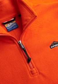 Sudadera de forro polar naranja con cuello de media cremallera, que presenta un tirador de cremallera negro y un parche con el logo. Textura suave y diseño liviano.
