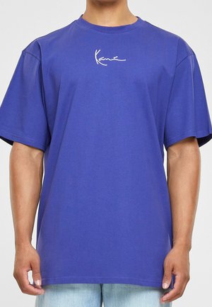 Person iført en enkel blå kortærmet t-shirt med en lille hvid signatur-stil logo på brystet, kombineret med lyseblå jeans.