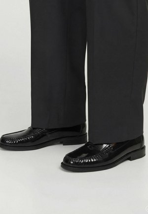 Crne polirane kožne mokasine nošene s crnim hlačama ravnih nogavica na jednostavnoj svijetloj pozadini.