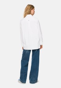 White long-sleeve shirt with a classic collar, paired with wide blue denim jeans featuring studded embellishments at the hem.