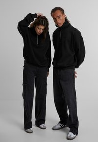 Svarte zip-up hettegensere med høy krage, kombinert med svarte cargo-bukser. Begge antrekkene har en avslappet passform og teksturert stoff.