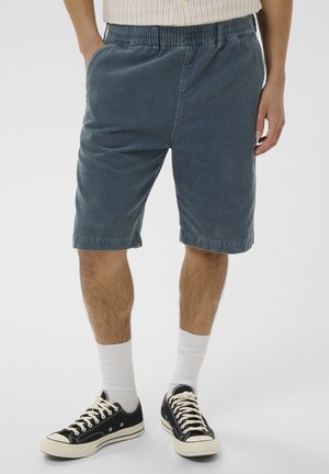 Mand iført blå kordfløjls-knælange shorts, hvide mellemlange sokker og sorte lave canvas-sneakers, stående mod en ensfarvet baggrund.