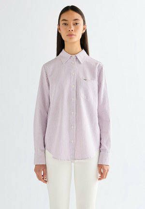 Kobieta ubrana w koszulę z pionowymi paskami w kolorze lawendy i bieli, z długimi rękawami, oraz białe spodnie, stojąca na jednolitym tle.