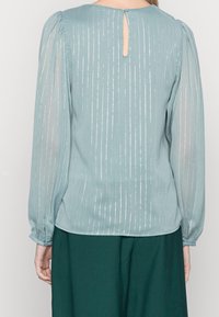 Blusa azzurro chiaro con strisce verticali argentate, maniche lunghe trasparenti, chiusura a bottoni sul retro e dettagli arricciati sulle spalle.