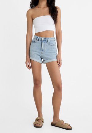 Mujer con un top corto sin mangas blanco, pantalones cortos de mezclilla azul claros con dobladillo enrollado y sandalias beige con hebilla, de pie sobre un fondo blanco.