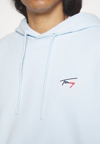 Sudadera con capucha azul claro hecha de tela suave, con un bolsillo tipo canguro, capucha con cordón y un pequeño logo bordado en rojo y azul marino.