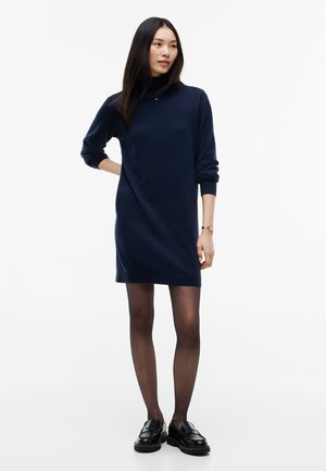 Kvinde iført en navyblå kjole med lange ærmer, sorte transparente strømpebukser, sorte loafers og et armbåndsur, stående med den ene hånd på hoften.
