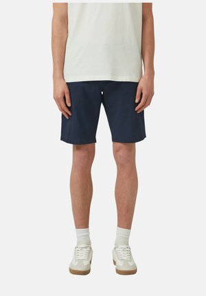 Ein Mann trägt marineblaue Shorts, weiße Socken, weiße Turnschuhe und ein helles Hemd und steht vor einem einfarbigen hellen Hintergrund.