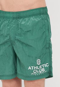 Zöld úszónadrág könnyű, gyűrött anyagból, rugalmas derékkal. Jellemzője a fehér "ATHLETIC CLUB" felirat és egy oldalsó zseb.