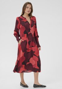 Langärmliges, rot-schwarzes Kleid mit V-Ausschnitt, gerafftem Taillenbereich und seitlichen Taschen. Glattes Material mit abstraktem Muster. Trägt schwarze Ballerinas.
