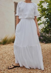 Vestido largo blanco y fluido con escote redondo, mangas cortas y bajo con volantes. Fabricado con tela ligera, con bolsillos laterales.