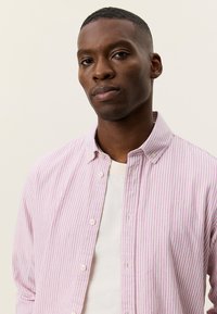 Ριγέ πουκάμισο με κουμπιά σε ροζ και λευκό ύφασμα, με κλασικό γιακά και τσέπη στο στήθος με ένα μικρό λογότυπο. Φοριέται πάνω από ένα λευκό t-shirt.