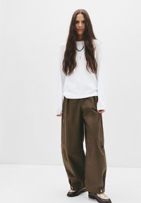 Long-sleeve white top paired with wide olive-green pants featuring zip pockets, and dark shoes with a textured design. Neutral background.