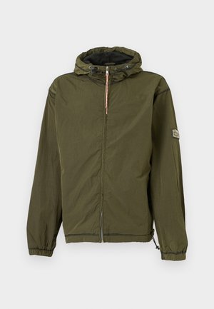 Hombre con chaqueta con capucha y cremallera verde oliva, camiseta blanca, pantalones de chándal negros descoloridos y zapatillas negras, de pie con las manos en los bolsillos.