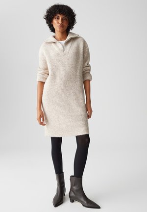 Gehaakte beige sweaterjurk met een ritskraag, geribbelde textuur en lange mouwen, gecombineerd met zwarte panty’s en enkelboots.