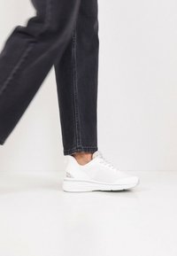 Λευκό αθλητικό παπούτσι με υφή στο πάνω μέρος, σχέδιο με κορδόνια και παχιά, μαλακή σόλα. Διακοσμητικά σε ανοιχτό γκρι τόνο κοντά στη φτέρνα.