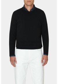 Schwarzer gestrickter Pullover mit rundem Halsausschnitt, langen Ärmeln und gerippten Bündchen, kombiniert mit weißen Hosen und einem blauen Button-Down-Hemd darunter.