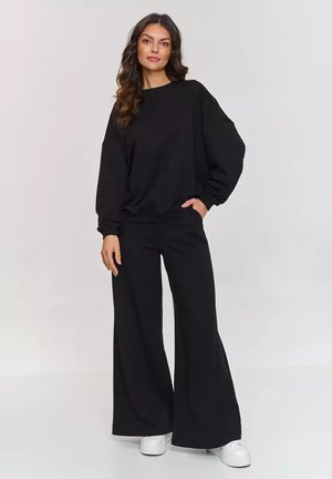 Czarna bluza z luźnym krojem i balonowymi rękawami, w połączeniu z szerokimi czarnymi spodniami. Model nosi białe sneakersy. Gładka tekstura.