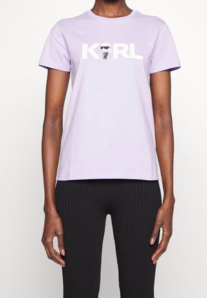 Osoba nosząca jasnofioletowy T-shirt z "KARL" i małą kreskówkową postacią w okularach przeciwsłonecznych nadrukowaną na biało, w połączeniu z czarnymi prążkowanymi spodniami.