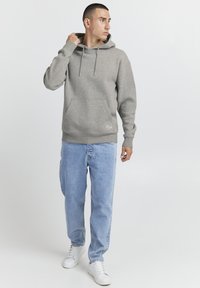 Sudadera gris hecha de tela suave, con un bolsillo frontal tipo canguro, cordones y puños acanalados. Combinada con jeans de color azul claro y zapatillas blancas.