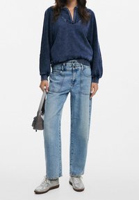 Pullover blu navy con colletto a mezza zip, abbinato a jeans dritti blu chiaro. Il modello tiene una piccola borsa grigia, indossando sneakers argentate.