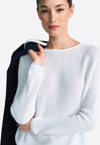 Biely pletený sveter s rebrovanou textúrou a dlhými rukávmi, doplnený tmavým sako prehodeným cez jedno rameno. Jednoduchý okrúhly výstrih.