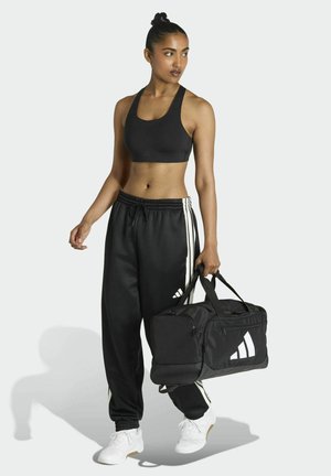 Top corto atletico nero, pantaloni neri larghi con righe laterali bianche e scarpe da ginnastica bianche. Porta una grande borsa da palestra nera con dettagli bianchi.