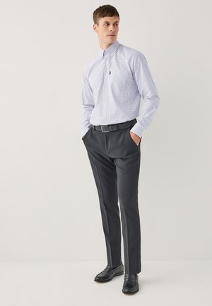 Uomo in piedi con le mani in tasca che indossa una camicia bianca a quadri, pantaloni neri, cintura nera e scarpe eleganti nere su uno sfondo semplice.