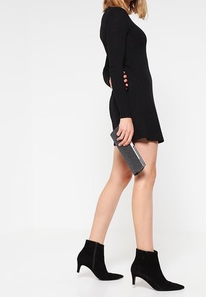 Kvinde i sort minikjole med lange ærmer og sorte ankelstøvler, der holder en grå clutch og står foran en hvid baggrund.