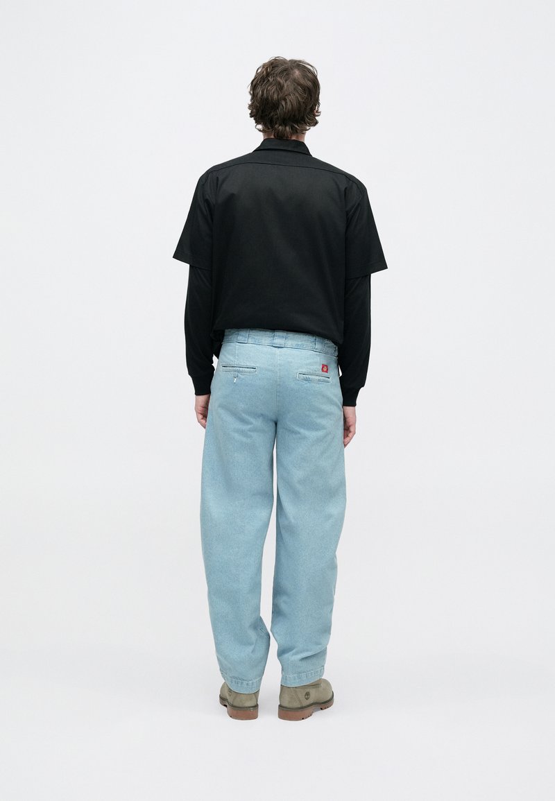 Chemise noire à manches courtes associée à un jean ample bleu clair. Le jean possède des poches arrière avec une étiquette rouge. Les chaussures sont de couleur foncée et plates.