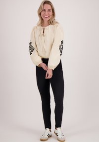 Crème blouse met een keyhole halslijn en zwarte geborduurde bloemenaccenten op de mouwen, gecombineerd met zwarte slim-fit broek en witte sneakers.