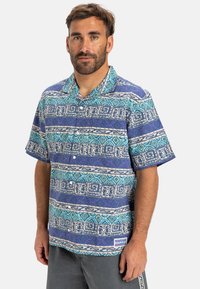 Camisa de botones de manga corta con un patrón geométrico en azul y morado, tela de algodón y una etiqueta en el dobladillo para la identificación de la marca.