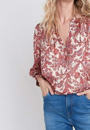 Blouse à motifs floraux rouge et crème avec un décolleté en V, des manches froncées et une texture douce. Associée à un jean en denim bleu.