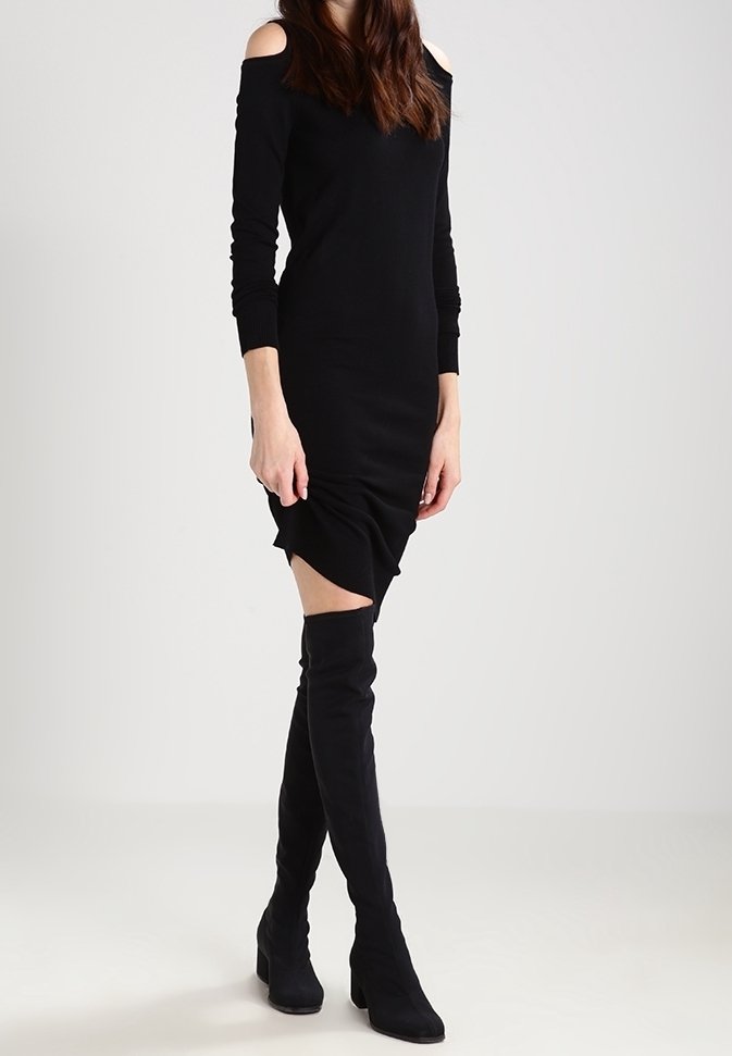Femme portant une robe moulante noire à épaules dénudées, associée à des bottes noires à talons montant au-dessus du genou, debout devant un fond clair uni.