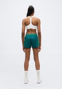 Γυναίκα που στέκεται με την πλάτη, φορώντας λευκό αθλητικό σουτιέν, πράσινο αθλητικό σορτς Under Armour, λευκές κάλτσες και λευκά αθλητικά παπούτσια σε απλό φόντο.