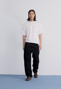 Camiseta blanca de gran tamaño, pantalones negros holgados y zapatos marrones. Diseño simple, sin patrones visibles, sobre un fondo claro.
