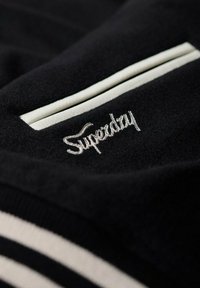 Svart fleecepullover med vit randdetalj och broderad "Superdry"-logotyp på bröstfickområdet. Mjukt material och avslappnad design.