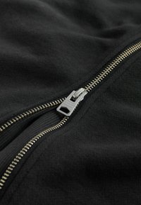 Melns džemperis ar rāvējslēdzēju un strukturētu audumu; tam ir sudrabota metāla rāvējslēdzēja aizdare un redzamas šuves gar atvēruma malu.