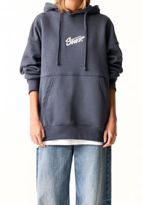 Tmavě šedá oversized mikina s kapucí, přední kapsou a bílým logem. Vyrobeno z měkkého materiálu s žebrovanými manžetami a lemem.