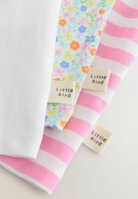 Tres prendas de bebé dobladas con etiquetas que dicen "Little Bird"; los patrones incluyen blanco con rayas rosas, flores en tonos pastel y blanco liso.