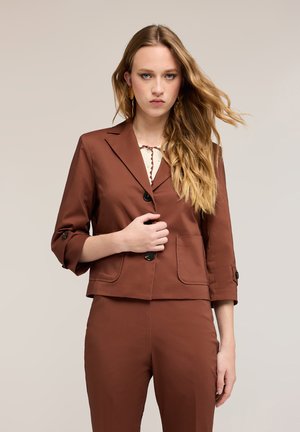 Nuori nainen, jolla on pitkät vaaleat hiukset, pukeutuneena ruskeaan napitettuun bleiseriin, jossa on kolmen neljänneksen hihat, ja matching housuihin yksinkertaisen taustan edessä.