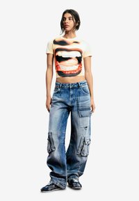 Krátke tričko s grafikou úst v červenej a čiernej farbe, v kombinácii s nadrozmernými modrými džínsovými cargo nohavicami s viacerými vreckami a zipsami.