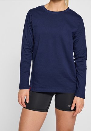 Femme portant un t-shirt à manches longues bleu marine et un short de sport noir ajusté, debout devant un fond clair uni.