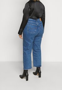 Vaqueros de mezclilla azul de cintura alta con corte recto; cuentan con dos bolsillos traseros y un parche de cuero con logo en la cintura. Se llevan con una blusa negra y botas de tacón.