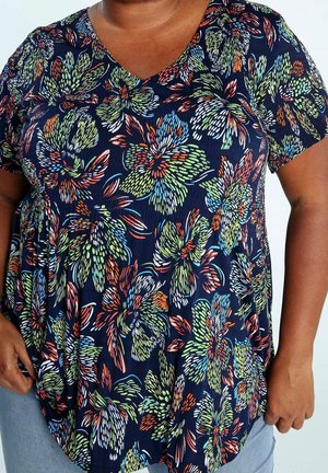 Person iført en mørkeblå skjorte med flerfarvede store blomsterprint og korte ærmer, kombineret med lyseblå jeans.