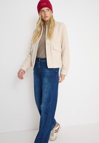 Crème fleece jack met voorzakken, gecombineerd met een lichtbruine top en donkerblauwe wijdleg jeans. Gemaakt met een rode rib gebreide beanie en sneakers.